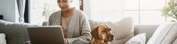 Woman using a laptop whilst sat on a sofa with a dog